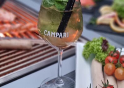 Campari Aperitif Glas mit Minze auf dem Grilltisch des Wirtshaus Alpinia, im Hintergrund frische Grillspezialitäten