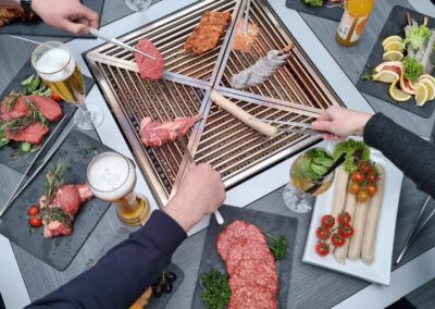 Gäste grillen gemeinsam am zentralen Grilltisch des Wirtshaus Alpinia mit Fleisch, Fisch und Garnelen