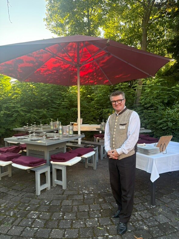 Servicemitarbeiter des Wirtshaus Alpinia am eingedeckten Grilltisch unter dem roten Sonnenschirm im Sommergarten