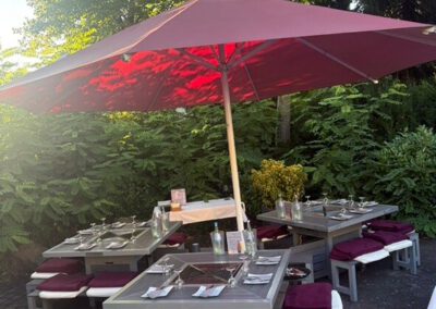Mehrere eingedeckte Grilltische mit Sitzbänken unter dem roten Sonnenschirm im Sommergarten des Wirtshaus Alpinia